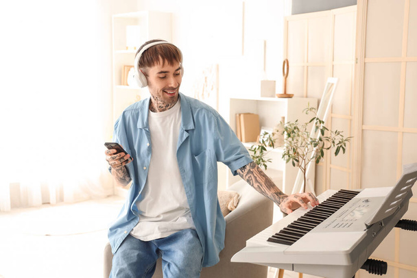 Joven hombre tatuado en auriculares con teléfono móvil jugando sintetizador en casa - Foto, Imagen