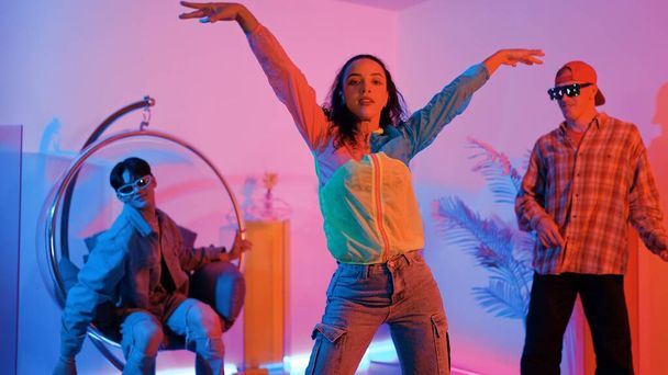 Ελκυστική ευτυχισμένη χορεύτρια κοιτάζοντας κάμερα, ενώ έξυπνη γυναίκα κινείται σε ομοιοκαταληξία με νέον φως. Επαγγελματικός ισπανόφωνος χορευτής break dancing φορώντας πολύχρωμο ύφασμα με ποικίλο φίλο. Ανάπλαση. - Φωτογραφία, εικόνα
