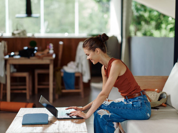Woman working on her laptop in a cozy home office The sunny summery background and vibrant tropical decor create a relaxing atmosphere for this freelancer With a big smile on her face, she - Photo, Image