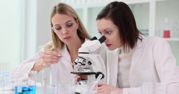 Az egyenruhás női tudós mikroszkóppal vizsgálja a mintát a laboratóriumban. A szemüveges szőke asszisztens reagenst ad a minta lassított felvételéhez. - Felvétel, videó