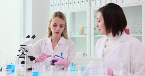 ブルネットの女性科学者は,実験室でブロンドのアシスタントに鮮やかな試薬を提供しています. 経験豊富な労働者が若い従業員を教える - 映像、動画