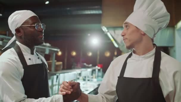 Молодой кавказский повар в фартуке и шляпе здоровается с чернокожим коллегой рукопожатием и разговаривает с ним на коммерческой кухне - Кадры, видео