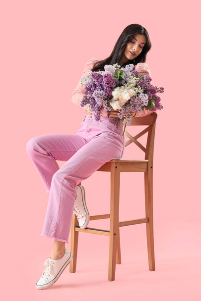 Beautiful young woman with lilac flowers sitting in chair on pink background - Photo, Image