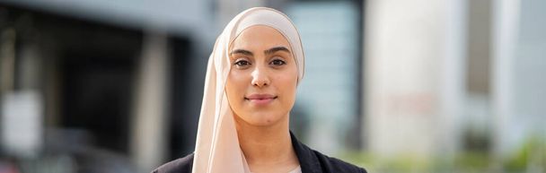 Heitere und doch selbstbewusste muslimische Geschäftsfrau im Hijab, die in einer modernen städtischen Landschaft steht und ihre berufliche Stärke und Eleganz widerspiegelt. - Foto, Bild