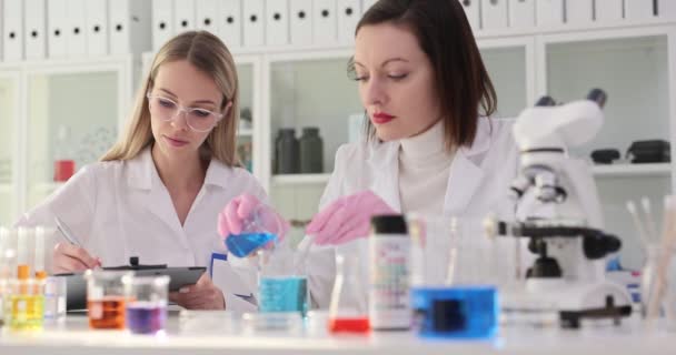 Vrouwelijke wetenschapper in handschoenen giet reagens in glas met vloeistof roeren met stok. Blondine assistent bereidt documentatie voor onderzoek - Video