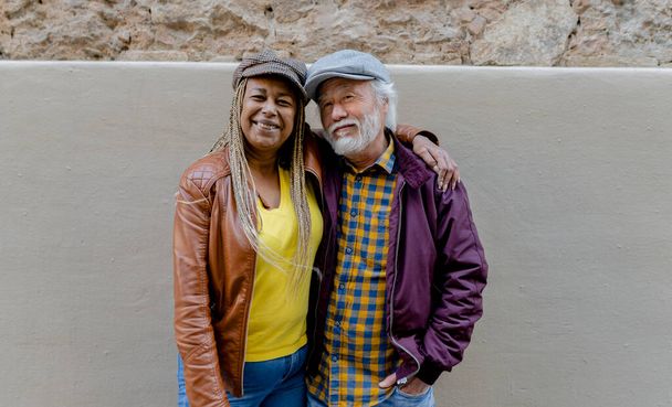 Okouzlující a různorodý seniorský pár se vřele usmívá a přitom se těsně objímají, zdůrazňují svou náklonnost a stylové městské oblečení na pozadí venkovské zdi. - Fotografie, Obrázek