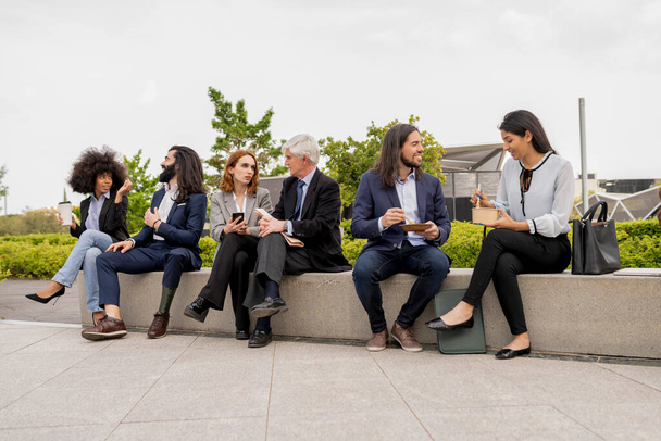 Professionelle Gruppe mit unterschiedlichem Hintergrund diskutiert Geschäftsstrategien während einer Kaffeepause auf einem Stadtplatz, was eine entspannte, aber produktive Arbeitskultur widerspiegelt. - Foto, Bild