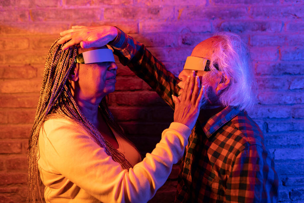 Пожилая пара исследует виртуальную реальность, руки трогают, окружены красочным освещением. - Фото, изображение