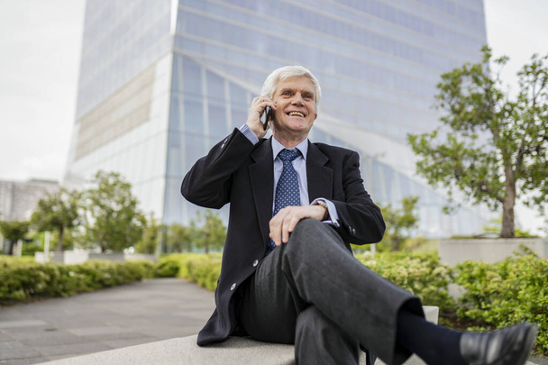 Sonriente hombre de negocios de edad avanzada en un traje haciendo una llamada telefónica mientras está sentado al aire libre. - Foto, Imagen