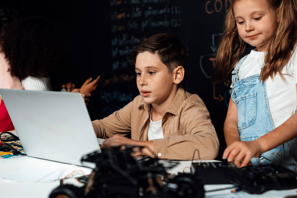 In classroom student in STEM class learning about coding robotics car. Brown shirt Schoolboy and white bib schoolgirl watching laptop and discussing. Other schoolgirls playing around funny. Erudition. - Photo, Image