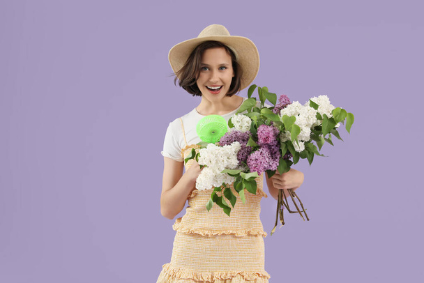 Jonge vrouw met groene kleine elektrische ventilator en boeket van lila bloemen op lila achtergrond - Foto, afbeelding