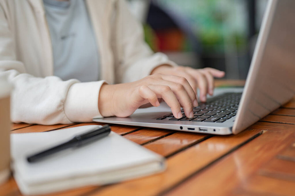 Uma imagem de close-up de uma mulher em uma camisola branca usando seu computador portátil, digitando no teclado do laptop, trabalhando remotamente em uma cafeteria. pessoas e conceitos de tecnologia sem fio - Foto, Imagem
