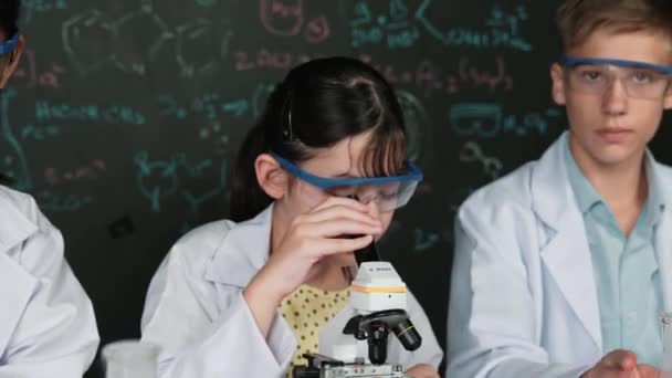 Menina bonito olhando sob microscópio enquanto estudante fazendo experiência em quadro-negro com teoria escrita. Jovem cientista inspeciona solução colorida à mesa com equipamento experimental colocado. Edificação - Filmagem, Vídeo