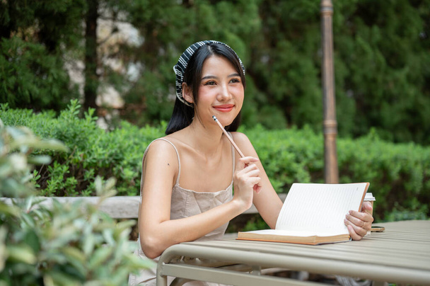 Una hermosa mujer asiática en un lindo vestido está soñando despierto mientras escribe su diario en el jardín verde patio trasero. personas y conceptos de ocio - Foto, Imagen