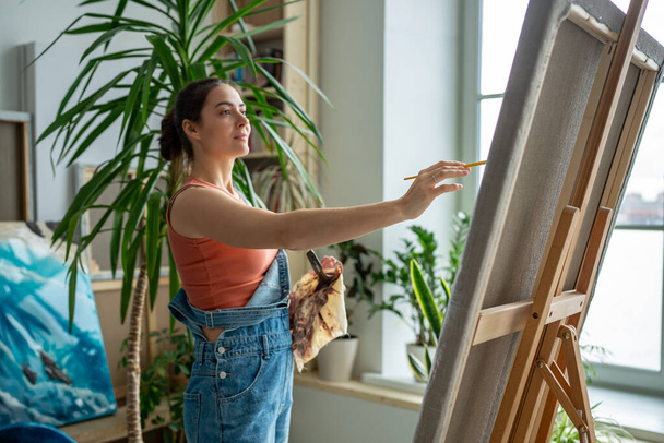 Happy smiling artistic female working on painting. Outstretched hand of satisfied glad woman with brush reaches for canvas, making careful thoughtful brushstrokes, creating masterpieces, artworks. - Photo, Image