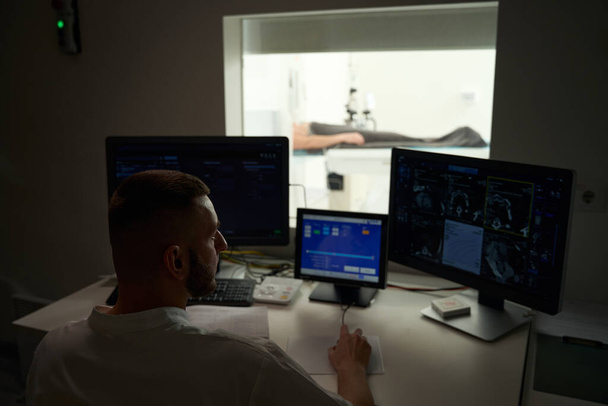コンピュータモニターで患者のCTスキャンを見るコントロールルームのデスクに座っている放射線技術者 - 写真・画像