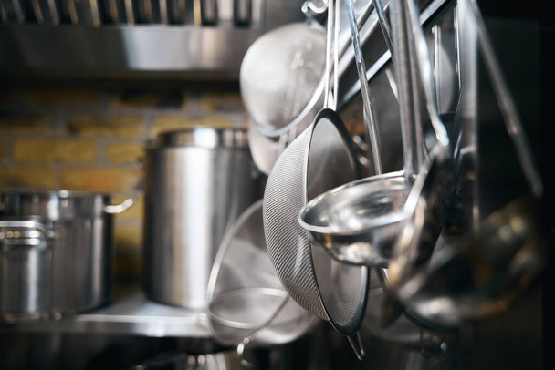 Focus on foreground of hanging cooking utensils and blurred table with saucepans on background in restaurant. Concept of tasty healthy eating - Photo, Image