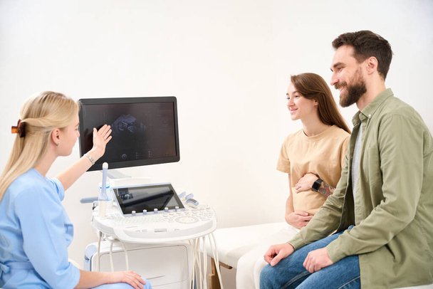 Női nőgyógyász mutatja képernyő eredményeként ultrahang szkennelés nő beteg terhes has fiatal mosolygós európai pár nézi orvos a klinikán. A terhesség és az apaság fogalma - Fotó, kép