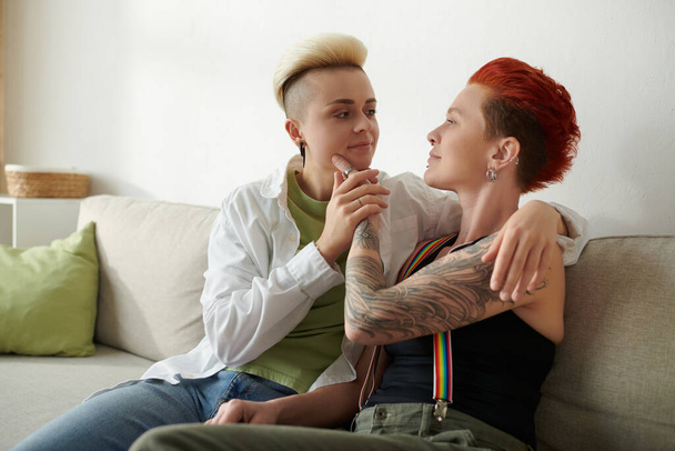 Dos mujeres con tatuajes se sientan juntas en un sofá, mostrando su arte corporal único en un ambiente interior acogedor. - Foto, Imagen