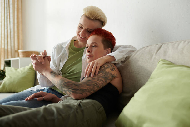 Dwie lgbt kobiety z tatuażami siedzieć wygodnie na kanapie, dzieląc chwilę wspólnoty i wyrażania siebie w domu. - Zdjęcie, obraz