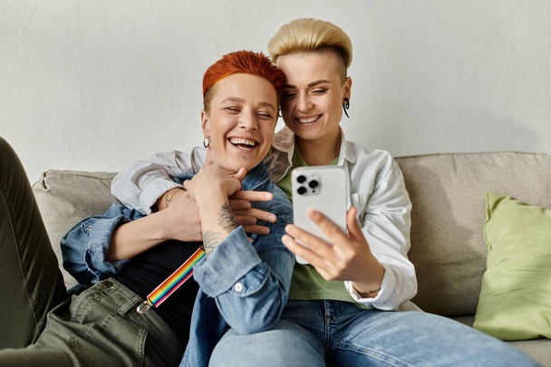 Egy rövid hajú leszbikus pár ül együtt a kanapén, mosolyognak és szelfit csinálnak egy telefonnal.. - Fotó, kép