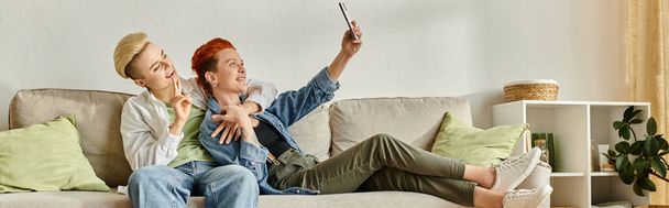 Ένας άνδρας και μια γυναίκα με κοντά μαλλιά κάθονται σε έναν καναπέ, χαμογελώντας και βγάζοντας μια selfie μαζί στην άνεση του σπιτιού τους. - Φωτογραφία, εικόνα