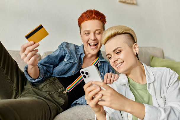Deux femmes, un couple lesbien aux cheveux courts, s'assoient sur un canapé tenant une carte de crédit, faisant du shopping en ligne ensemble à la maison. - Photo, image
