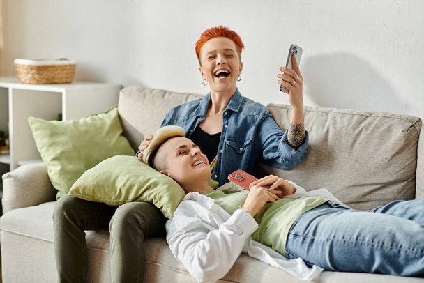 Deux femmes, un couple lesbien aux cheveux courts, s'assoient étroitement sur un canapé, absorbées dans leur téléphone portable. - Photo, image