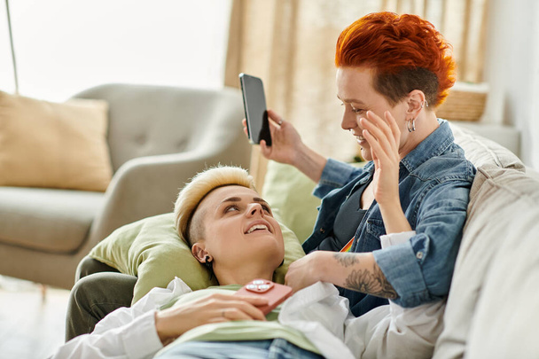 Dos mujeres con el pelo corto participan en una conversación mientras están sentadas de cerca en un sofá en casa. - Foto, Imagen