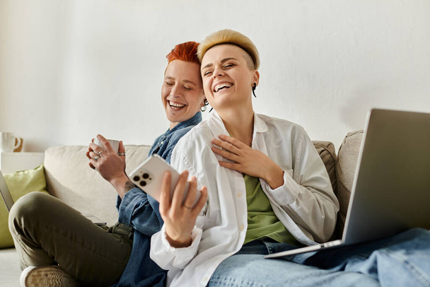 Duas mulheres, um casal lésbico com cabelo curto, sentam-se em um sofá, focadas em seus laptops em casa.. - Foto, Imagem