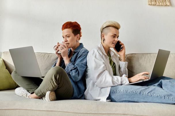Lesbijska para z krótkimi włosami siedzi na kanapie, każda pochłonięta własnym laptopem. - Zdjęcie, obraz