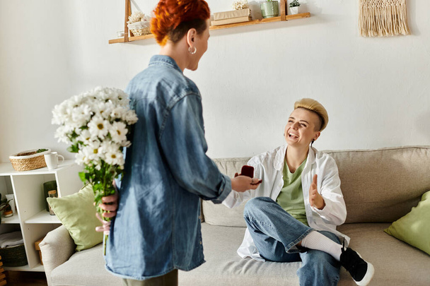 Una donna con i capelli corti presenta un mazzo di fiori al suo partner, che è seduto su un divano. Home setting, gesto romantico. - Foto, immagini