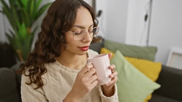 Μια στοχαστική νεαρή γυναίκα με γυαλιά πίνοντας μια κούπα καφέ σε ένα άνετο σαλόνι. - Πλάνα, βίντεο