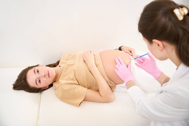 Ginecóloga que se inyecta con jeringa en el vientre embarazada de una joven europea que mira a la cámara en la clínica. Concepto de embarazo y maternidad - Foto, imagen
