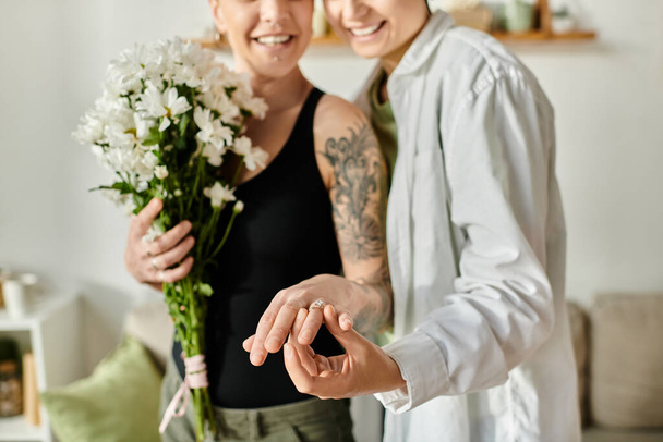 Két nő virágcsokrot tart egy hangulatos nappaliban, eljegyzési gyűrűt mutatva be. - Fotó, kép