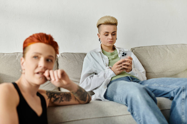 Δύο άνθρωποι, ένα ζευγάρι λεσβιών με κοντά μαλλιά, κάθονται σε έναν καναπέ απορροφημένο από το τηλέφωνο, αποκομμένοι ο ένας από τον άλλο. - Φωτογραφία, εικόνα