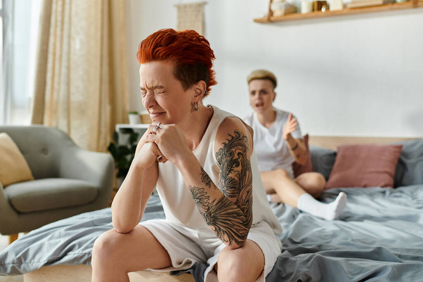 αναστατωμένος γυναίκα με τατουάζ κάθονται μαζί με το σύντροφό και κλαίνε σε ένα κρεβάτι σε ένα υπνοδωμάτιο, αναδεικνύοντας τη μοναδική τέχνη του σώματος τους. - Φωτογραφία, εικόνα