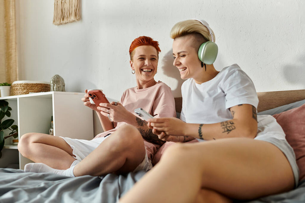 Δύο γυναίκες με κοντά μαλλιά κάθονται σε ένα κρεβάτι, ασχολούνται με το να παίζουν παιχνίδια μαζί, επιδεικνύοντας ένα σύγχρονο τρόπο ζωής ΛΟΑΤ. - Φωτογραφία, εικόνα