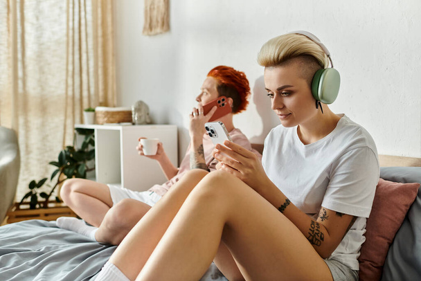 Ένα λεσβιακό ζευγάρι με κοντά μαλλιά κάθεται σε ένα κρεβάτι, απορροφημένο με μουσική μέσω ακουστικών, ενσωματώνοντας την ουσία του ΛΟΑΤ τρόπου ζωής. - Φωτογραφία, εικόνα
