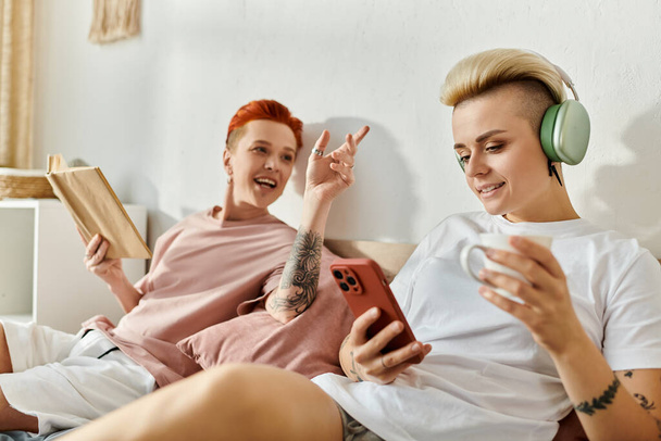 Para lesbijek z krótkimi włosami siedzi na łóżku, nosząc słuchawki i ciesząc się muzyką w sypialni. - Zdjęcie, obraz