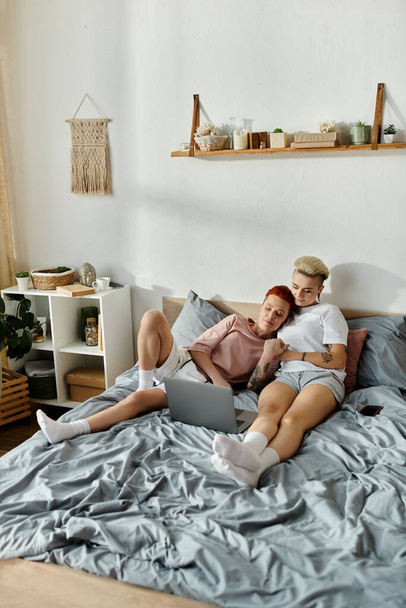 Ένα ζευγάρι λεσβιών με κοντά μαλλιά κάθεται σε ένα κρεβάτι, βυθισμένο στην οθόνη του φορητού υπολογιστή τους, επιδεικνύοντας ένα σύγχρονο τρόπο ζωής ΛΟΑΤ. - Φωτογραφία, εικόνα