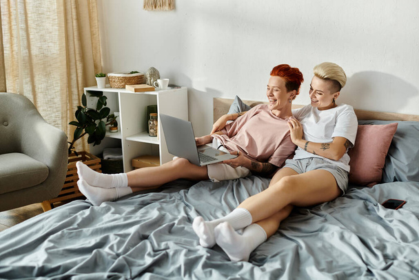 Δύο γυναίκες με κοντά μαλλιά κάθονται σε ένα κρεβάτι, απορροφημένες χρησιμοποιώντας ένα φορητό υπολογιστή. - Φωτογραφία, εικόνα