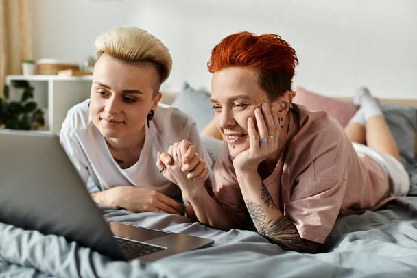 Δύο άνθρωποι, ένα ζευγάρι λεσβιών με κοντά μαλλιά, χαλαρώστε και συνδεθείτε σε ένα κρεβάτι, ενώ κοιτάζοντας μια οθόνη laptop. - Φωτογραφία, εικόνα