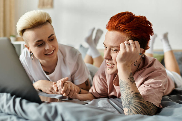 Δύο γυναίκες με κοντά μαλλιά ξαπλώνουν σε ένα κρεβάτι, απορροφημένες σε μια οθόνη laptop, μοιράζονται μια ζεστή και οικεία στιγμή. - Φωτογραφία, εικόνα