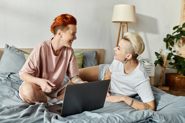 Δύο γυναίκες με κοντά μαλλιά κάθονται σε ένα κρεβάτι, απορροφημένες σε μια οθόνη laptop, αγκαλιάζοντας τον ψηφιακό κόσμο μαζί. - Φωτογραφία, εικόνα