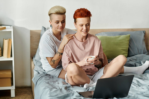 Deux femmes aux cheveux courts s'assoient sur un lit, absorbées dans un écran d'ordinateur portable. - Photo, image
