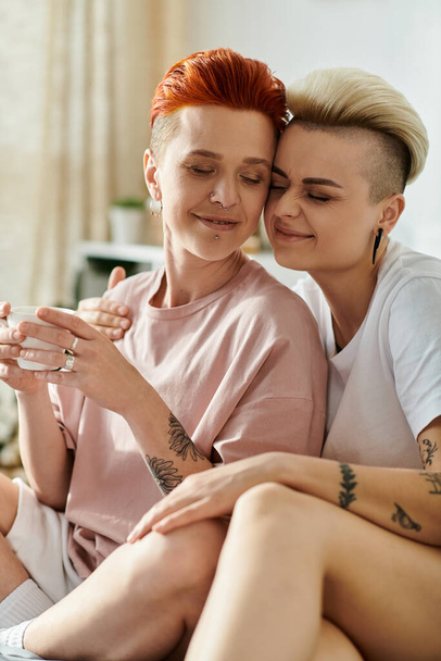 Δύο γυναίκες με τατουάζ, η μία με κοντό κούρεμα, κάθονται σε ένα κρεβάτι σε μια κρεβατοκάμαρα, επιδεικνύοντας τα μοναδικά τους τατουάζ.. - Φωτογραφία, εικόνα