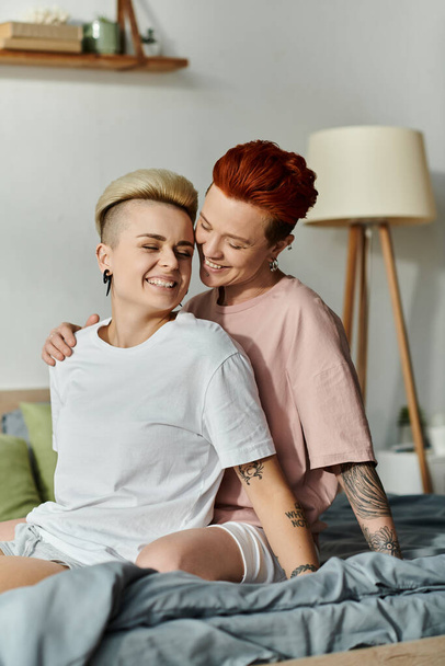 Теплый момент, когда двое людей, лесбийская пара с короткими волосами, сидят на кровати и обнимаются в своей спальне.. - Фото, изображение