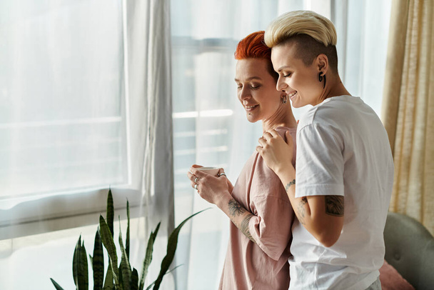Two women, a lesbian couple with short hair, stand embracing in front of a window in their bedroom, showcasing their happy LGBT lifestyle. - Photo, Image