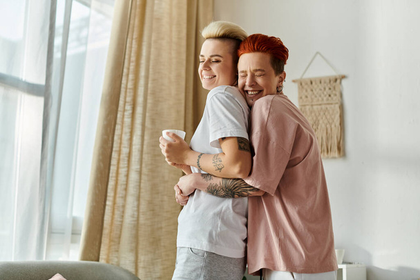 Лесбийская пара с короткими волосами нежно обнимается перед окном, демонстрируя свою любовь и близость. - Фото, изображение